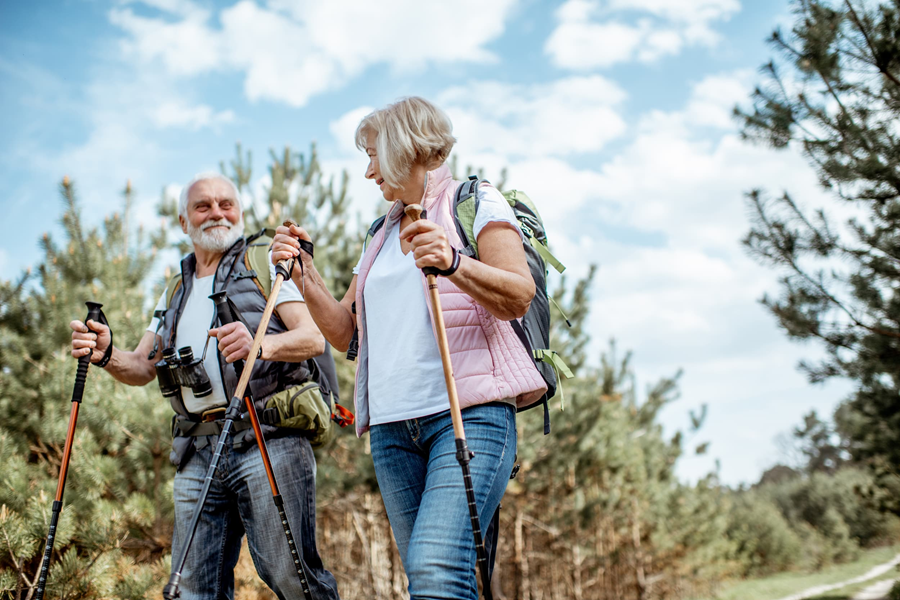 A couple in their seventies use trekking sticks to hike through a wooded area.